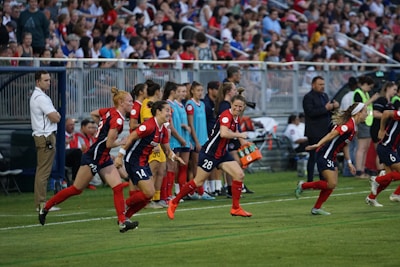 A group of female soccer players in red and blue uniforms are actively running towards the field, indicating a substitution or start of the game. The scene is vibrant with many spectators in the background, sitting on bleachers and watching the event. There are coaching staff and team members on the sidelines, along with sports equipment such as water coolers.