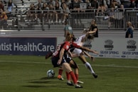 Two soccer players are engaged in action on a grass field with a partially filled stadium in the background. One player in red and black is positioned defensively, while the other player in white and yellow appears to be maneuvering around them. A blue and white soccer ball is visible near their feet. Several spectators can be seen seated in the stands.
