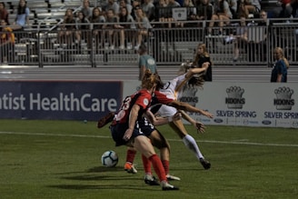 Two soccer players are engaged in action on a grass field with a partially filled stadium in the background. One player in red and black is positioned defensively, while the other player in white and yellow appears to be maneuvering around them. A blue and white soccer ball is visible near their feet. Several spectators can be seen seated in the stands.