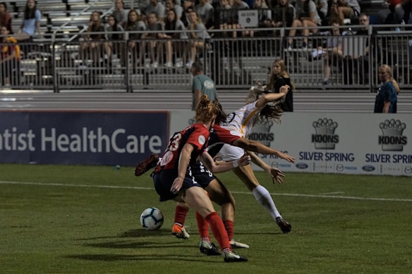 Two soccer players are engaged in action on a grass field with a partially filled stadium in the background. One player in red and black is positioned defensively, while the other player in white and yellow appears to be maneuvering around them. A blue and white soccer ball is visible near their feet. Several spectators can be seen seated in the stands.