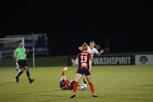 Several soccer players are engaged in an intense game on a field at night. One player in a red and blue uniform, marked with the number 23, is standing and observing the action. Another player is on the ground, wearing red shorts. A referee in a green jersey and black shorts is nearby. The background features a sign for 'WASHSPIRIT' and a partially visible goalpost.