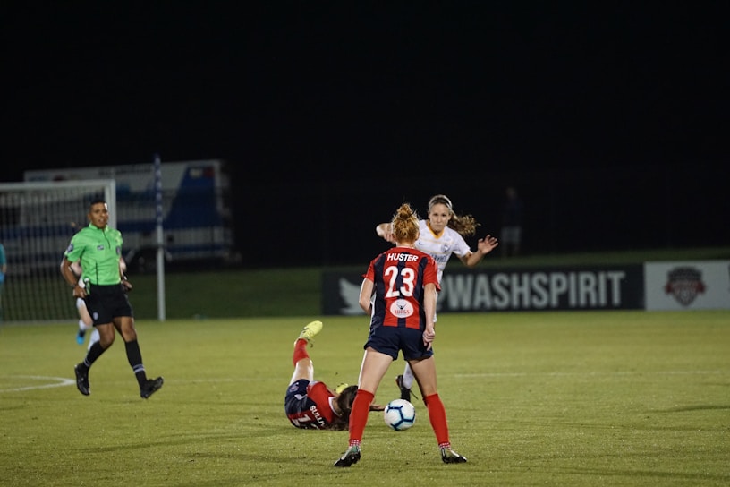 Several soccer players are engaged in an intense game on a field at night. One player in a red and blue uniform, marked with the number 23, is standing and observing the action. Another player is on the ground, wearing red shorts. A referee in a green jersey and black shorts is nearby. The background features a sign for 'WASHSPIRIT' and a partially visible goalpost.