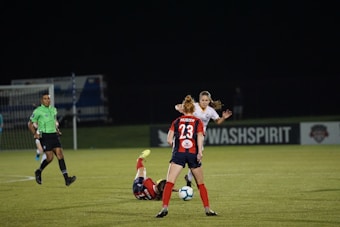 Several soccer players are engaged in an intense game on a field at night. One player in a red and blue uniform, marked with the number 23, is standing and observing the action. Another player is on the ground, wearing red shorts. A referee in a green jersey and black shorts is nearby. The background features a sign for 'WASHSPIRIT' and a partially visible goalpost.