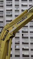 A yellow construction excavator arm with branding, positioned in front of a high-rise building with numerous windows.
