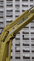 A yellow construction excavator arm with branding, positioned in front of a high-rise building with numerous windows.