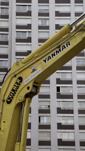 A yellow construction excavator arm with branding, positioned in front of a high-rise building with numerous windows.