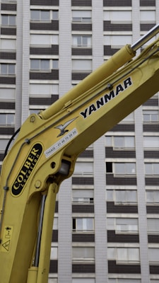 A yellow construction excavator arm with branding, positioned in front of a high-rise building with numerous windows.