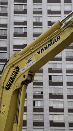 A yellow construction excavator arm with branding, positioned in front of a high-rise building with numerous windows.