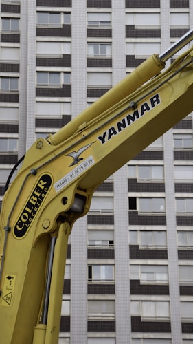 A yellow construction excavator arm with branding, positioned in front of a high-rise building with numerous windows.