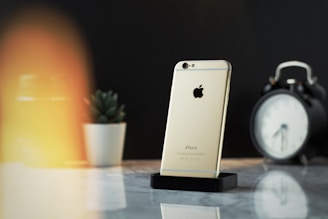 A gold iPhone is positioned upright on a black stand, set against a marble surface. In the background, there is a blurred white ceramic pot containing a small succulent plant, and an analog clock with a black casing. The lighting creates a warm glow on the left side of the image.