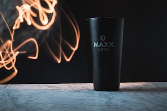 A black coffee tumbler with the words 'MAXX Coffee' printed on it is placed on a marbled surface. There are abstract streaks of warm light appearing in the background, creating a dynamic and creative atmosphere.