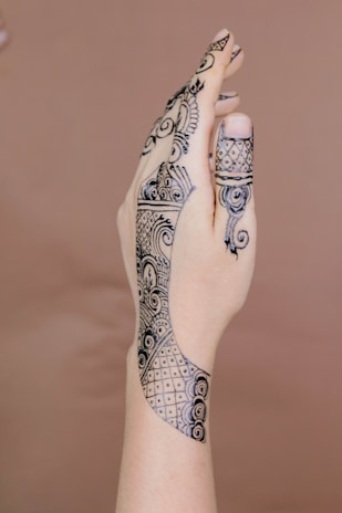 A hand adorned with intricate henna designs featuring a mix of floral and geometric patterns. The artwork extends from the fingertips, with dense details, and continues along the hand's back towards the wrist. The background is a plain, soft brown color, which helps highlight the black henna designs.