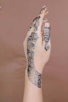 A hand adorned with intricate henna designs featuring a mix of floral and geometric patterns. The artwork extends from the fingertips, with dense details, and continues along the hand's back towards the wrist. The background is a plain, soft brown color, which helps highlight the black henna designs.