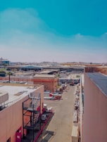A bustling industrial area with various buildings and structures, including warehouses and storage facilities. The street is lined with items and equipment, and a truck is parked in the distance. The scene is set under a clear blue sky.