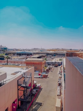 A bustling industrial area with various buildings and structures, including warehouses and storage facilities. The street is lined with items and equipment, and a truck is parked in the distance. The scene is set under a clear blue sky.