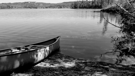 A black and white photograph of a calm lake with a canoe resting on the rocky shore in the foreground. Surrounding the lake are dense forests and distant hills. The water reflects the trees and sky, creating a tranquil scene.