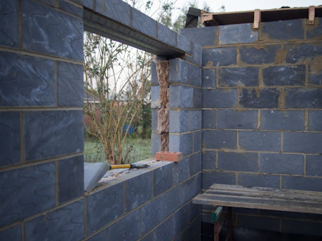 A partially constructed building with exposed bricks and an unfinished window opening. Through the window, vegetation and trees are visible outside. There are construction tools like a trowel and a level resting on the window ledge, and a wooden shelf inside.