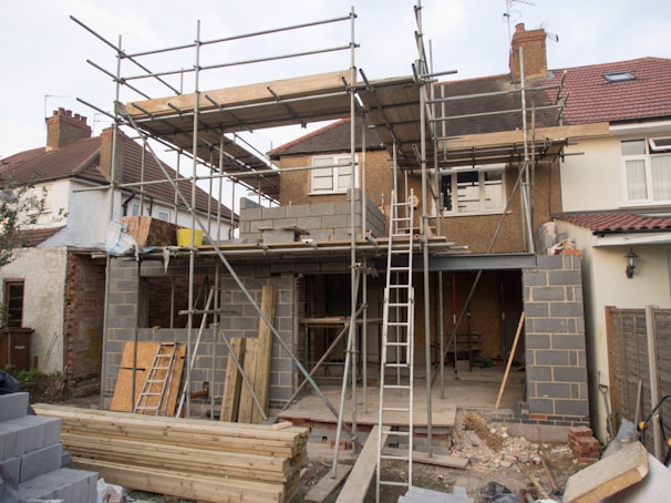 A house under construction with scaffolding surrounding the building. The structure includes wooden planks and cinder blocks. There are ladders leaning against the house, and various construction materials such as panels and timber are scattered around the site.