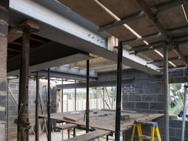 An under-construction building site with metal beams, scaffolding, and unfinished brick walls. A wooden plank is suspended between two structures, and construction materials are scattered across the area. A ladder is positioned against one of the walls.