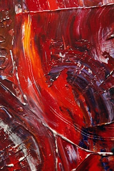 Vibrant red and orange abstract strokes layered with textures and curves create a dynamic visual flow across the canvas, accented by darker hues and subtle hints of white. The paint appears to have been applied thickly, giving a sense of movement and energy.