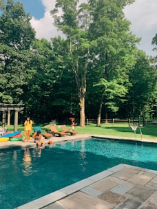 A swimming pool is surrounded by a landscaped backyard area with lush green grass and tall trees. There are lounge chairs and inflatable pool toys near the pool. People are sitting by the poolside, enjoying a sunny day. A basketball hoop is positioned by the pool.