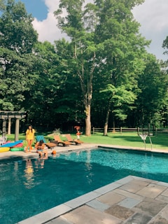 A swimming pool is surrounded by a landscaped backyard area with lush green grass and tall trees. There are lounge chairs and inflatable pool toys near the pool. People are sitting by the poolside, enjoying a sunny day. A basketball hoop is positioned by the pool.