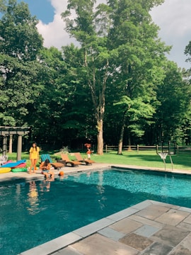 A swimming pool is surrounded by a landscaped backyard area with lush green grass and tall trees. There are lounge chairs and inflatable pool toys near the pool. People are sitting by the poolside, enjoying a sunny day. A basketball hoop is positioned by the pool.