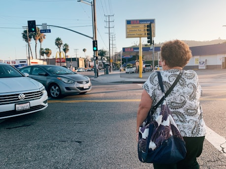 A middle-aged woman with short hair, wearing a patterned shirt and carrying a bag, is crossing a city street. Cars wait at the intersection, and palm trees are visible in the background. A gas station with price signs is on the corner, and the sun casts long shadows, indicating either early morning or late afternoon.
