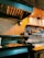 A commercial kitchen with a countertop holding several food items, including bread and stacked metal containers. The grill has some food cooking on it, likely a pizza or flatbread with toppings. Shelves above have large containers and other cooking supplies.
