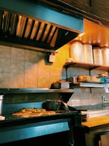 A commercial kitchen with a countertop holding several food items, including bread and stacked metal containers. The grill has some food cooking on it, likely a pizza or flatbread with toppings. Shelves above have large containers and other cooking supplies.