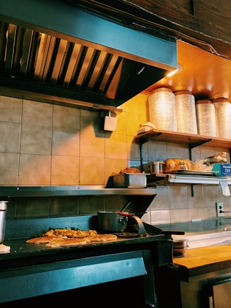 A commercial kitchen with a countertop holding several food items, including bread and stacked metal containers. The grill has some food cooking on it, likely a pizza or flatbread with toppings. Shelves above have large containers and other cooking supplies.