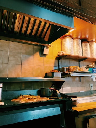 A commercial kitchen with a countertop holding several food items, including bread and stacked metal containers. The grill has some food cooking on it, likely a pizza or flatbread with toppings. Shelves above have large containers and other cooking supplies.