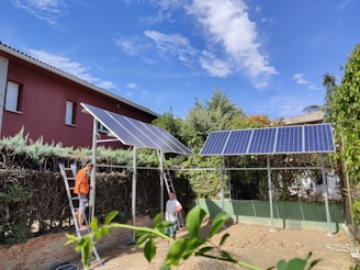 Two people are working with solar panels in a garden surrounded by vegetation. One person stands on a ladder installing or inspecting the panels. The area is bordered by tall hedges and trees, and a red house is visible in the background under a clear blue sky.
