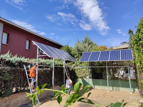 Two people are working with solar panels in a garden surrounded by vegetation. One person stands on a ladder installing or inspecting the panels. The area is bordered by tall hedges and trees, and a red house is visible in the background under a clear blue sky.