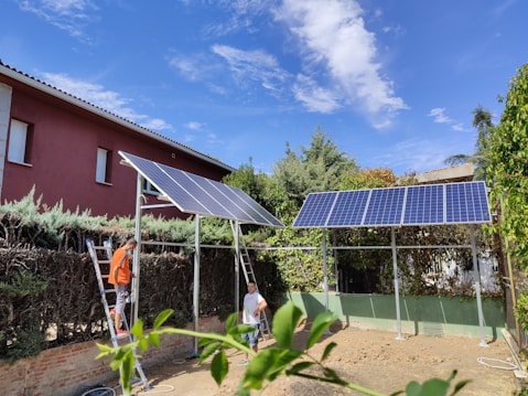 Two people are working with solar panels in a garden surrounded by vegetation. One person stands on a ladder installing or inspecting the panels. The area is bordered by tall hedges and trees, and a red house is visible in the background under a clear blue sky.