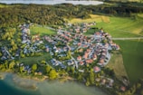 Aerial view of a small village surrounded by lush green fields and forests. The village is composed of numerous houses with red rooftops, arranged in a circular pattern. Roads interconnect the buildings, and large patches of vegetation and trees are interspersed between the structures. A body of water is visible in the background, adding to the tranquil rural setting.