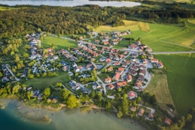 Aerial view of a small village surrounded by lush green fields and forests. The village is composed of numerous houses with red rooftops, arranged in a circular pattern. Roads interconnect the buildings, and large patches of vegetation and trees are interspersed between the structures. A body of water is visible in the background, adding to the tranquil rural setting.