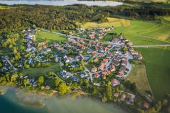 Aerial view of a small village surrounded by lush green fields and forests. The village is composed of numerous houses with red rooftops, arranged in a circular pattern. Roads interconnect the buildings, and large patches of vegetation and trees are interspersed between the structures. A body of water is visible in the background, adding to the tranquil rural setting.