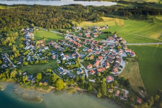 Aerial view of a small village surrounded by lush green fields and forests. The village is composed of numerous houses with red rooftops, arranged in a circular pattern. Roads interconnect the buildings, and large patches of vegetation and trees are interspersed between the structures. A body of water is visible in the background, adding to the tranquil rural setting.