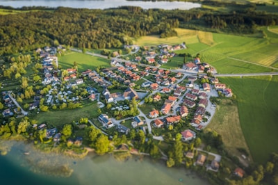 Aerial view of a small village surrounded by lush green fields and forests. The village is composed of numerous houses with red rooftops, arranged in a circular pattern. Roads interconnect the buildings, and large patches of vegetation and trees are interspersed between the structures. A body of water is visible in the background, adding to the tranquil rural setting.