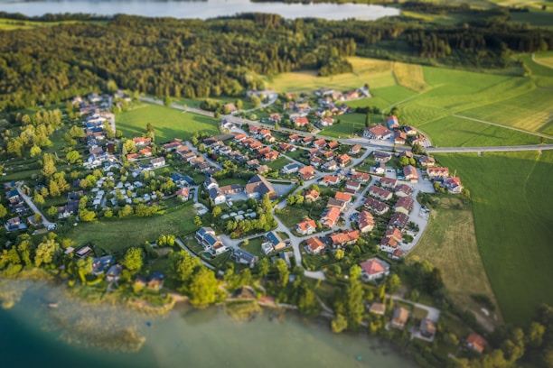 Aerial view of a small village surrounded by lush green fields and forests. The village is composed of numerous houses with red rooftops, arranged in a circular pattern. Roads interconnect the buildings, and large patches of vegetation and trees are interspersed between the structures. A body of water is visible in the background, adding to the tranquil rural setting.