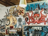 The image features a vibrant mural on a cafe wall with eclectic artwork, including a humanoid figure with a flaming head and a motorcycle, alongside a cartoonish octopus wearing a Route 66 top hat. There are abstract and text elements, including words like 'BREWS' and 'Route 66'. Below the mural, a coffee machine and takeaway cups are visible, adding a cafe-like atmosphere.