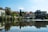A scenic riverside view with traditional Italian architecture lining the waterfront. The buildings have rustic designs, surrounded by lush green trees. A stone bridge crosses the river, which reflects the skyline beautifully. Bright daylight enhances the serene and picturesque setting.