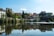 A scenic riverside view with traditional Italian architecture lining the waterfront. The buildings have rustic designs, surrounded by lush green trees. A stone bridge crosses the river, which reflects the skyline beautifully. Bright daylight enhances the serene and picturesque setting.