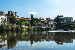 A scenic riverside view with traditional Italian architecture lining the waterfront. The buildings have rustic designs, surrounded by lush green trees. A stone bridge crosses the river, which reflects the skyline beautifully. Bright daylight enhances the serene and picturesque setting.