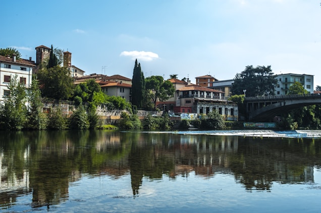 A scenic riverside view with traditional Italian architecture lining the waterfront. The buildings have rustic designs, surrounded by lush green trees. A stone bridge crosses the river, which reflects the skyline beautifully. Bright daylight enhances the serene and picturesque setting.