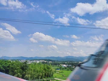 A high-speed train traveling through a lush, green landscape with a backdrop of distant mountains under a clear blue sky with scattered clouds. The foreground is slightly blurred, indicating the rapid motion of the train.