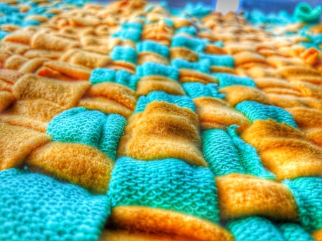 A close-up of a textured fabric with a woven pattern, featuring a combination of yellow and teal colored fibers. The fabric appears soft and plush, with a detailed view of both smooth and textured surfaces.
