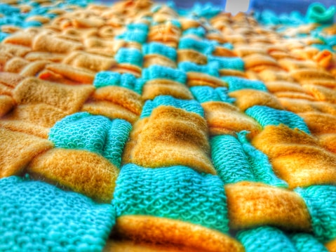 A close-up of a textured fabric with a woven pattern, featuring a combination of yellow and teal colored fibers. The fabric appears soft and plush, with a detailed view of both smooth and textured surfaces.