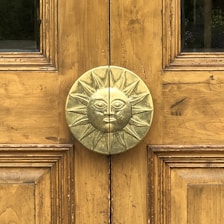 A wooden door features a central, ornate metallic doorknob in the shape of a sun with a face. The wood has a natural golden brown finish with visible grain patterns. The sun motif is detailed with rays extending outward, and the door is symmetrically paneled.